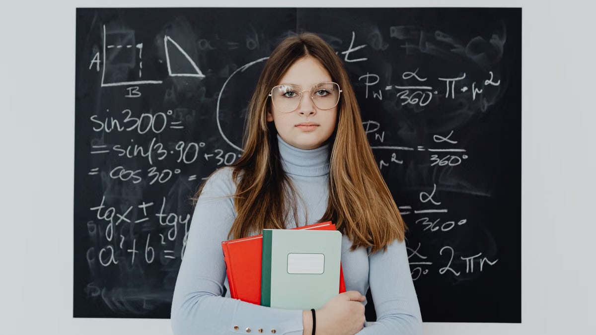 A teenage girl with glasses stands in front of a chalkboard, holding textbooks.